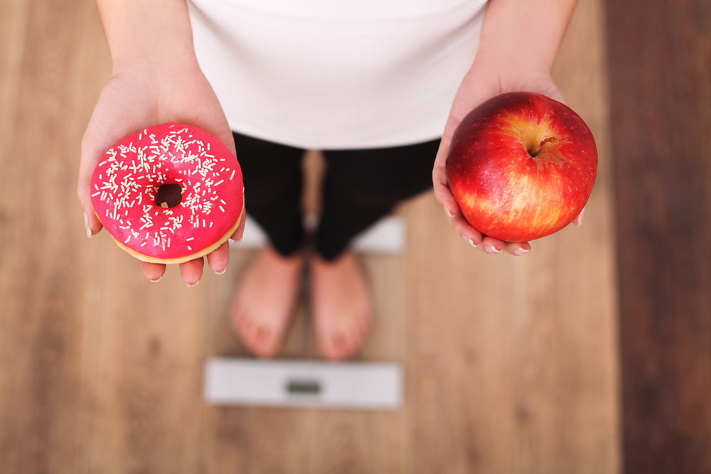 Frau hält Apfel in der einen und Donut in der anderen Hand während sie auf der Wage steht